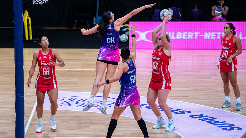 Players from Lightning and Forest, compete near the goalpost during a game. A player in red (Goal Shooter) prepares to shoot while a tall defender in purple (Goal Keeper) jumps to block the shot. Other players are positioned around the shooting circle, watching the play. The court branding and a digital screen in the background display 