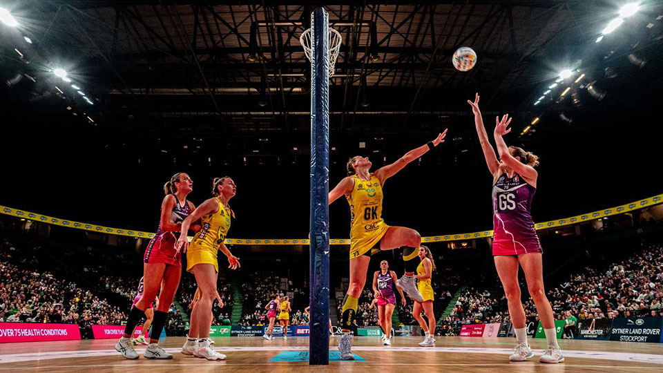 A wide-angle low shot of a Loughborough Lightning netball player shooting while Manchester Thunders reach to block. Photography by Ben Lumley