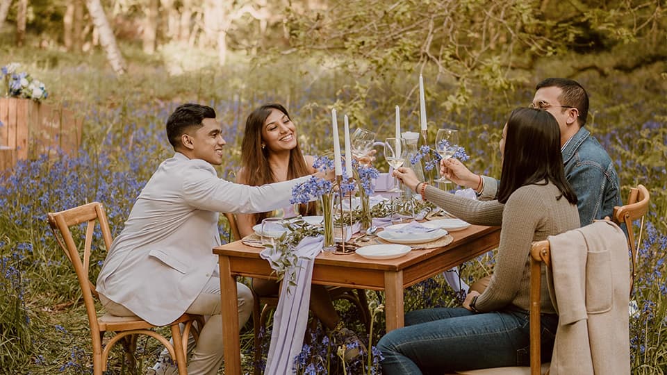 Aditya Mukherjee and Aarushi Mittal sitting at a wooden table with their friends surrounded by bluebells at Burleigh Wood, 麻豆視頻_麻豆直播_麻豆传媒官网. They are smiling and raising their glasses.
