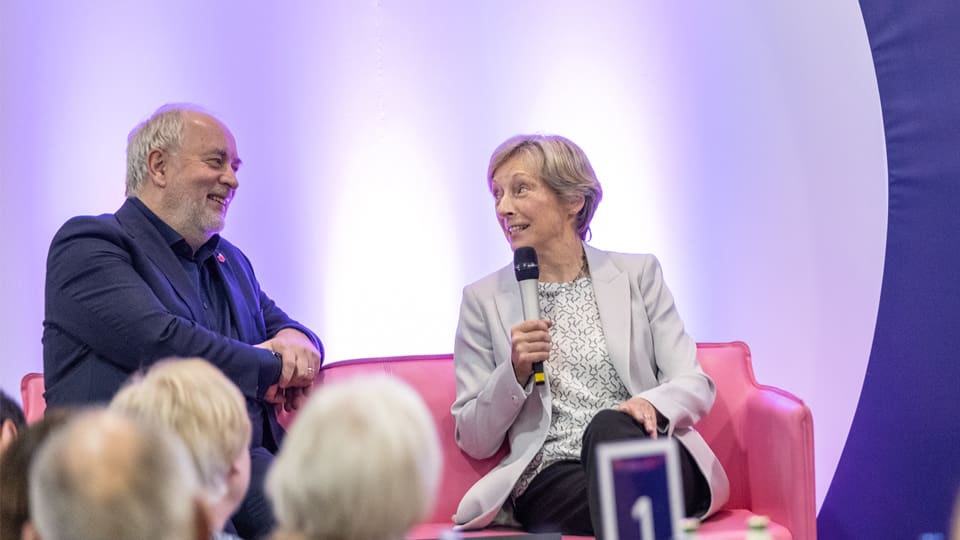 Professor Nick Jennings and Dame Liz Nicholl seated on a pink sofa. Liz holds a microphones and they are smiling.