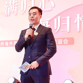 Winston is wearing a black suit with a pocket chief. He is holding a microphones and speaking and a bit of card looking out at an audience.