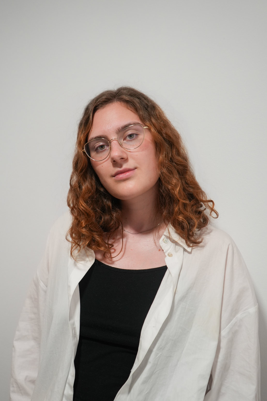 Kate, a female student with long brown hair and round glasses smiles at the camera, she is wearing a black t-shirt and white shirt