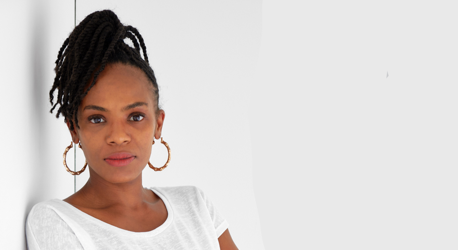 Kedisha Coakley a black woman in her late 30s with long black hair tied up in a white tshirt and large hoop earrings, stands against a white wall