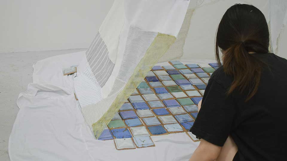 Art installation of ceramic tiles on a white floor with cloth hanging from above and a student with black hair and a black t-shirt sitting on the floor pictured from behind