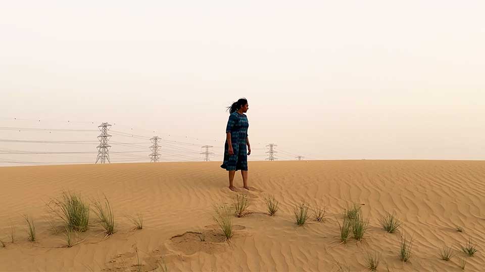 Young female wearing a long dress with dark hair tied back in a desert or dunes with electricity pylons visible in the background