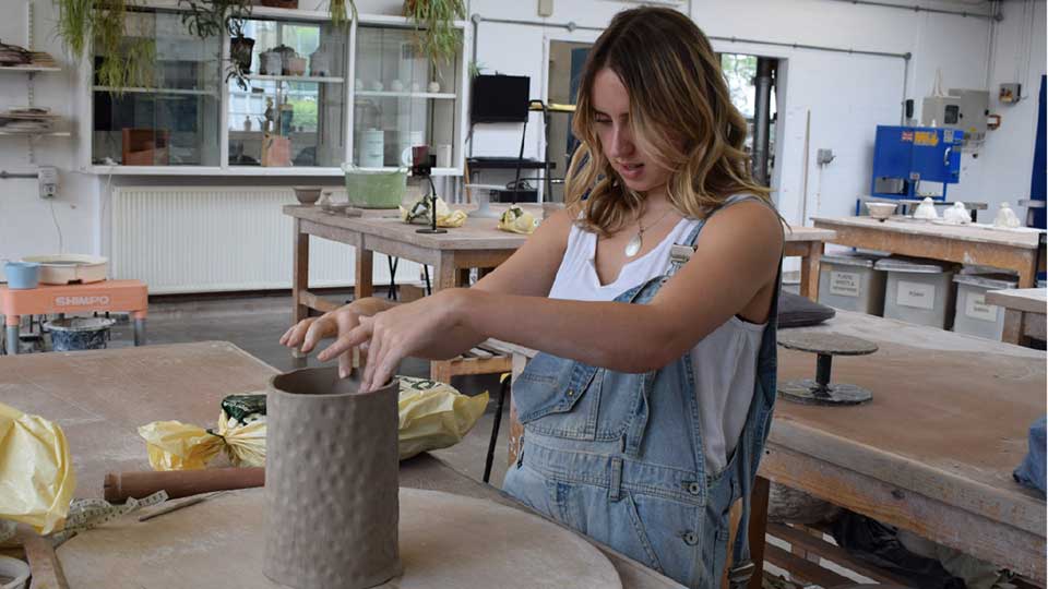 Female student in early twenties working on a large ornamental clay work in a ceramics workshop, wearing denim dungarees and a white top