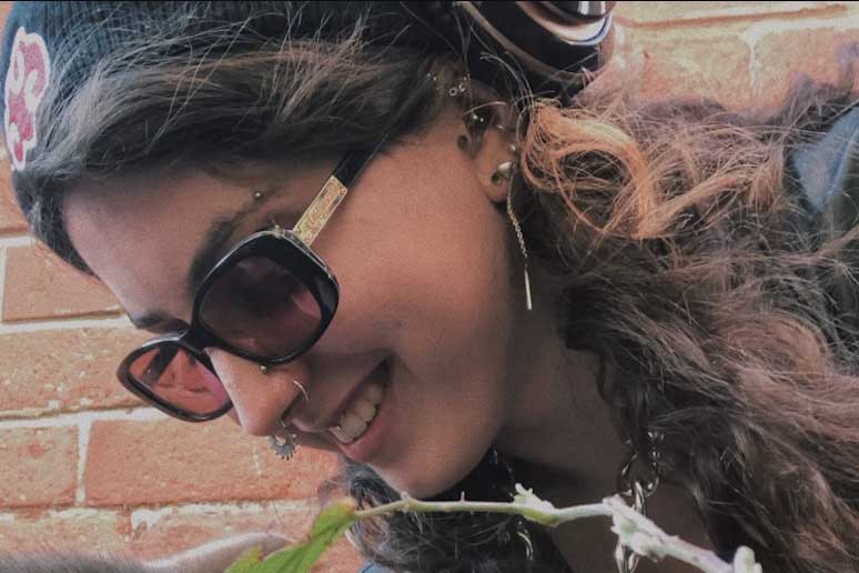 Headshot taken at a diagonal angle of a young female looking down and away from the camera. She is smiling, wearing a black knitted hat, over the ear headphoness (just visible), black sunglasses and has long wavy brown hair. She has multiple nose and ear piercings with a stud and rings.