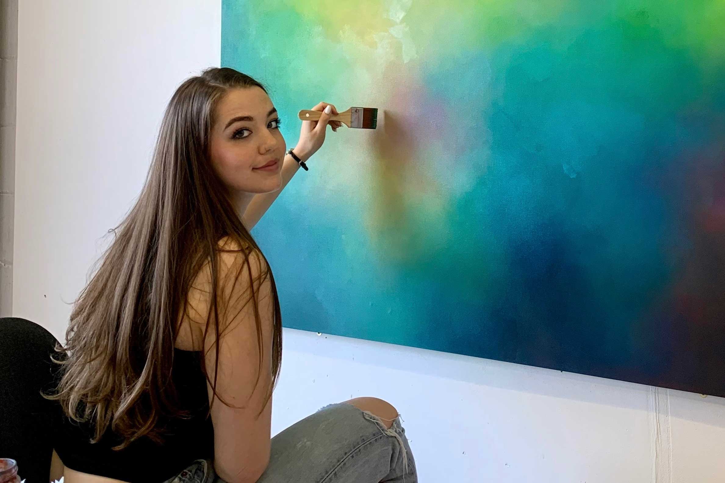 Young white female student, seated on an office style chair looking slightly over her shoulder at the camera while working on a large abstract brightly coloured painting of blues and greens. She has very long dark brown hair and is wearing a black vest top and ripped jeans.