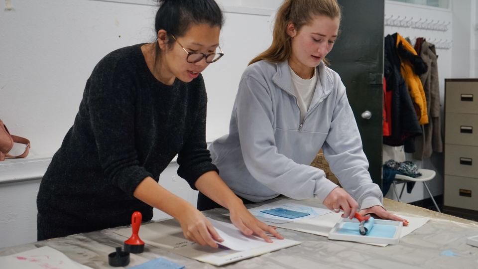 A photograph of two people standing rolling light blue paint onto paper