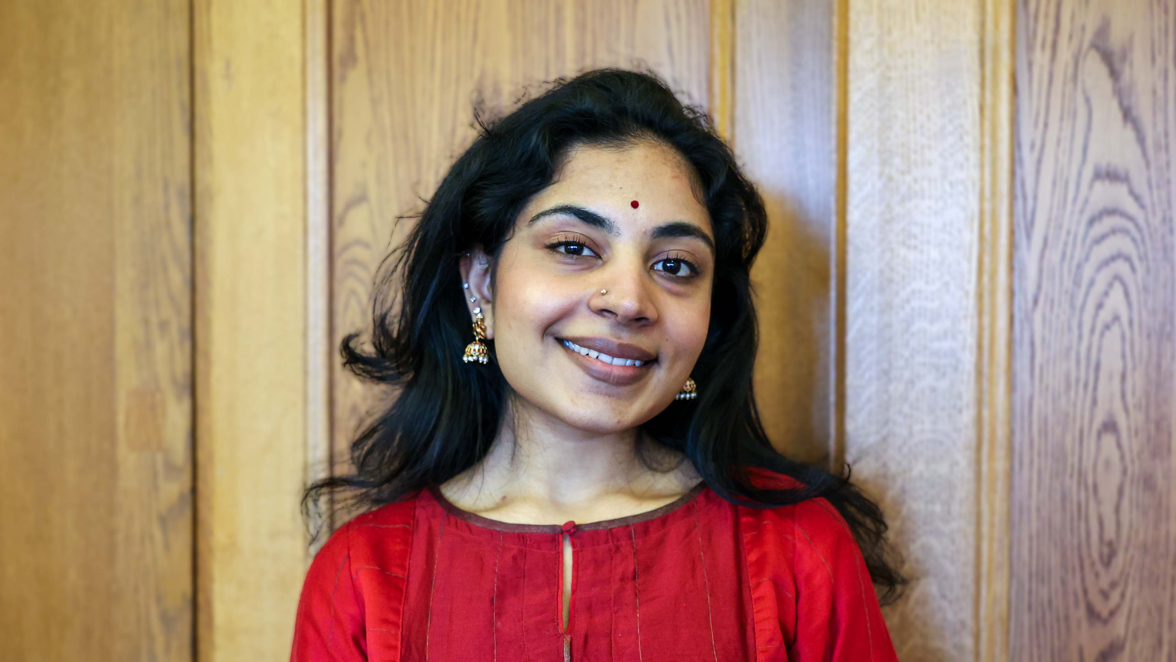 Young female from a south asian background with long black hair, wearing dark red and gold earrings and necklace