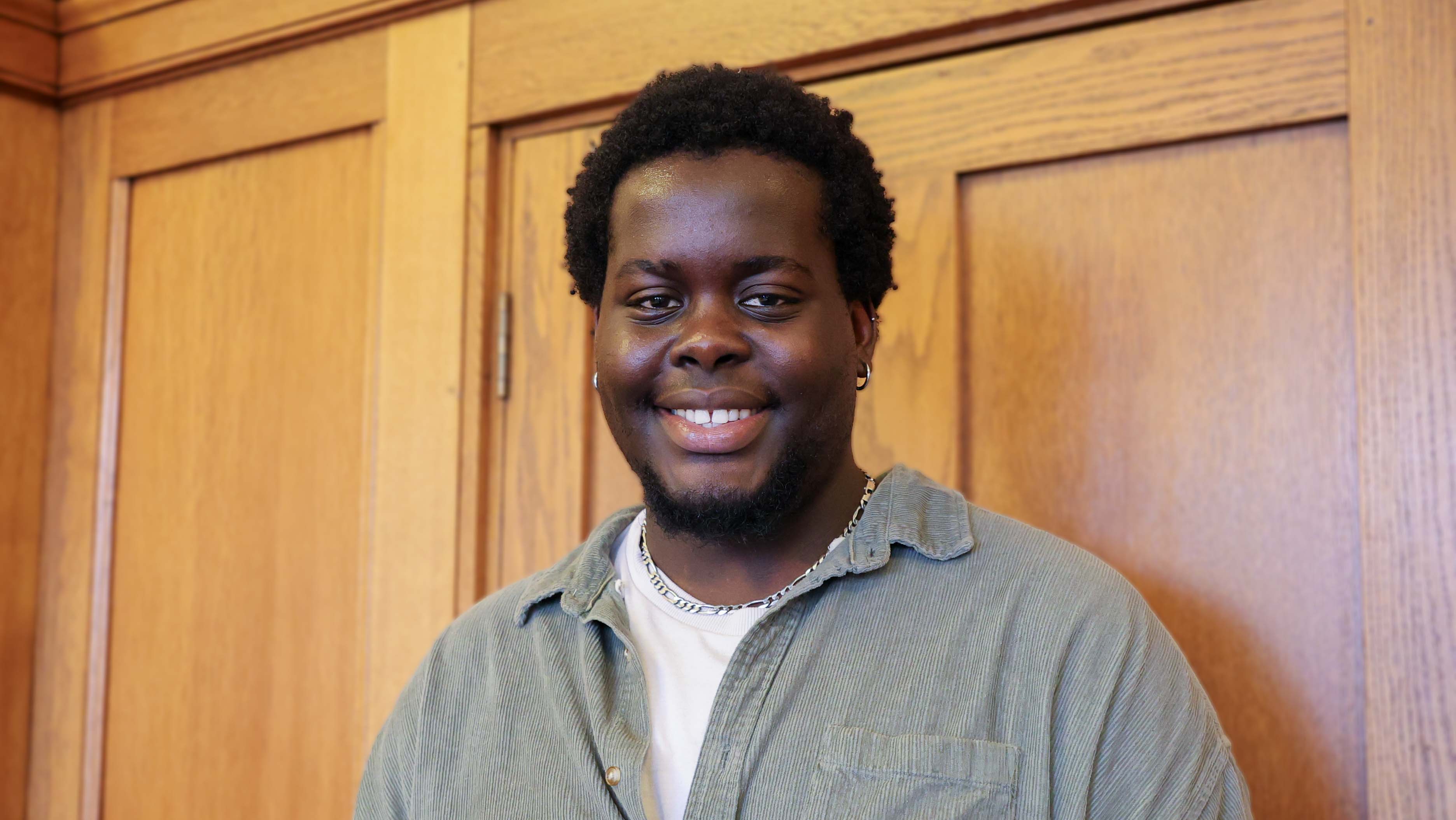 Young black male with short dark hair, wearing a grey shirt with a white t-shirt visible underneath and thick silver chain