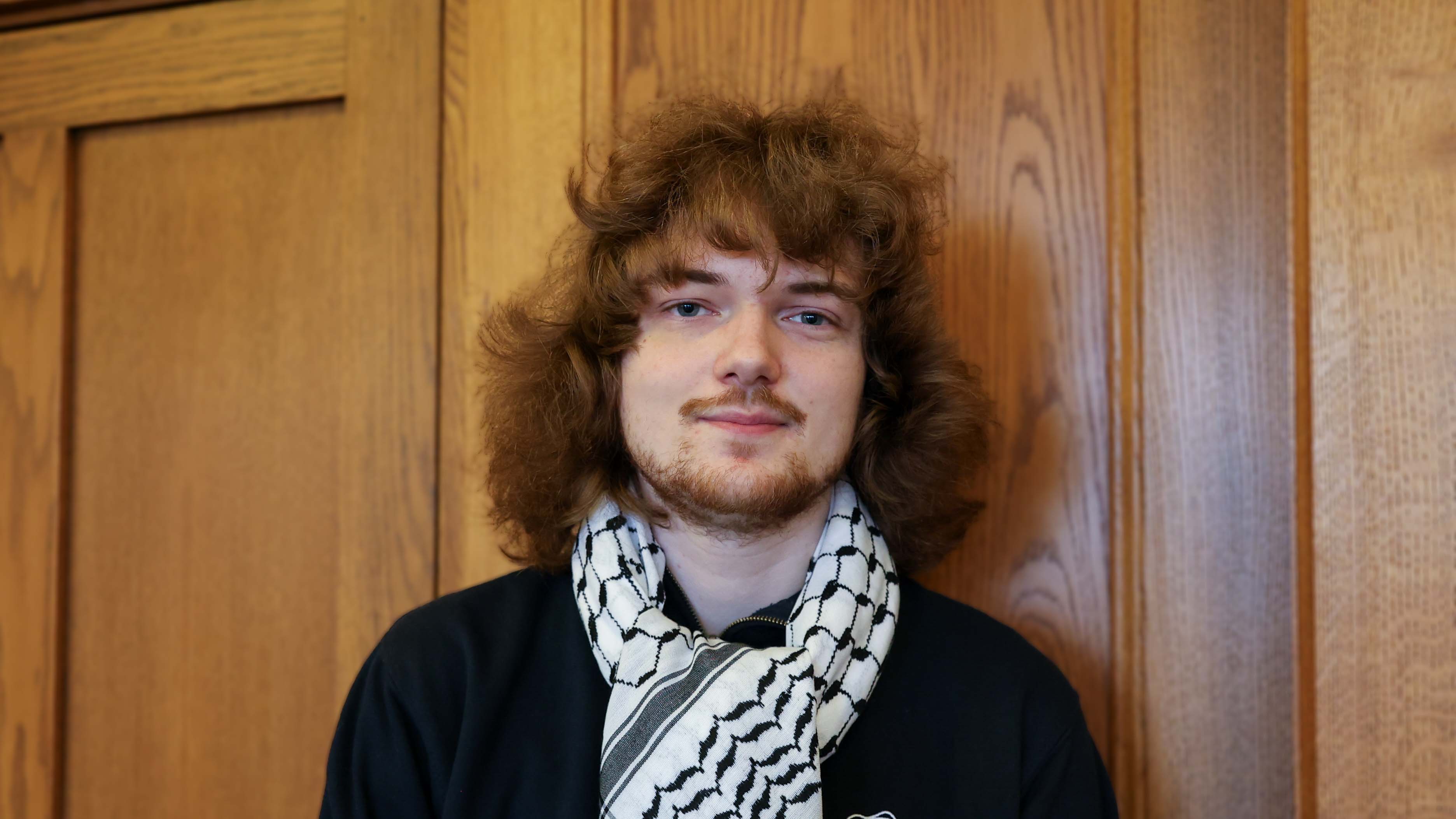 Young white male with shoulder length thick, wavy brown hair and facial hair wearing a black top and a black and white scarf
