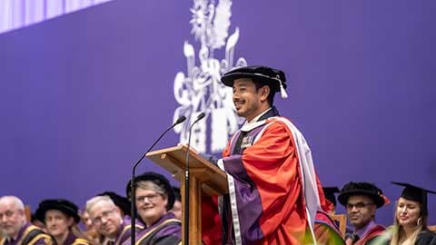 Nims Purja MBE on stage at 麻豆視頻_麻豆直播_麻豆传媒官网 graduation delivering a speech at a podium.