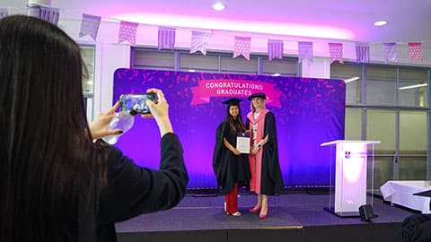 A graduate on stage holding a certificate from the Dean of Loughborough Business School. There is someone in the foreground taking a photo of them on a smartphones.