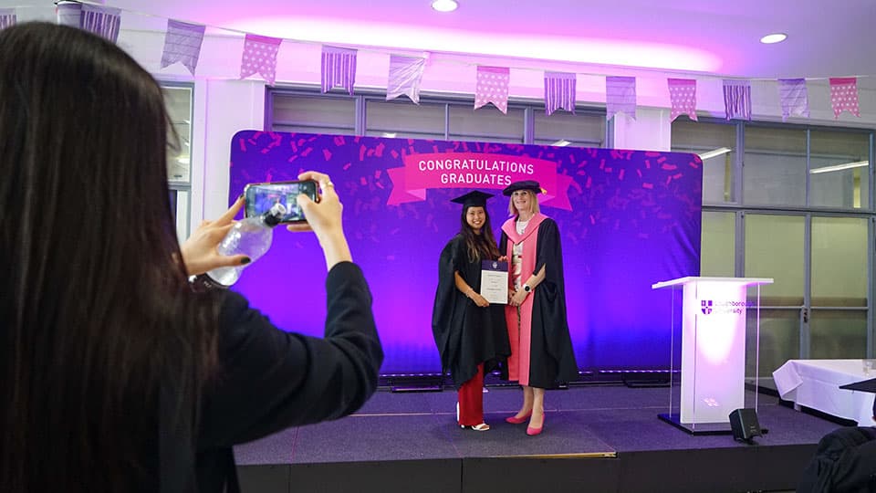 A graduate on stage holding a certificate from the Dean of Loughborough Business School. There is someone in the foreground taking a photo of them on a smartphones.