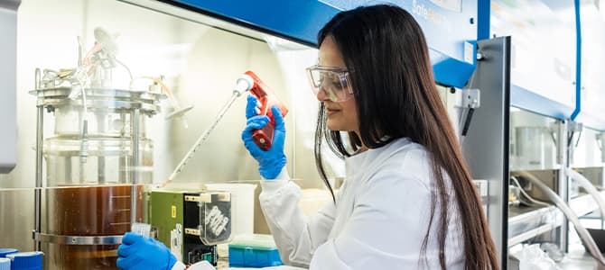 researcher wearing lab goggles, coat and gloves holding a test tube