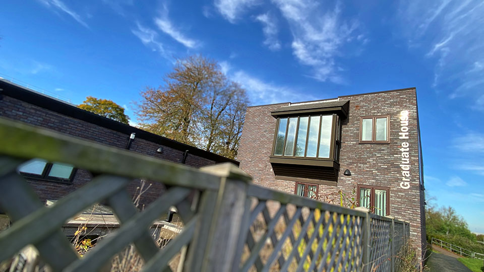 An oblique architectural view of Graduate House in sunshine and blue skies, displaying the vertical name signage in brushed metal at one corner.