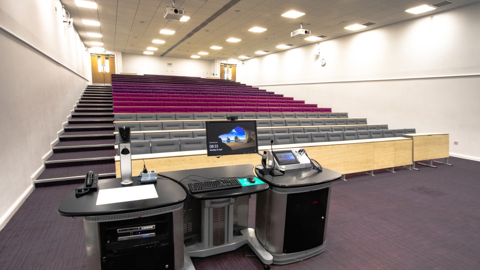James France CC011 - Lecture hall with tiered seating, purple and gray chairs, and a central podium equipped with computer and microphones.