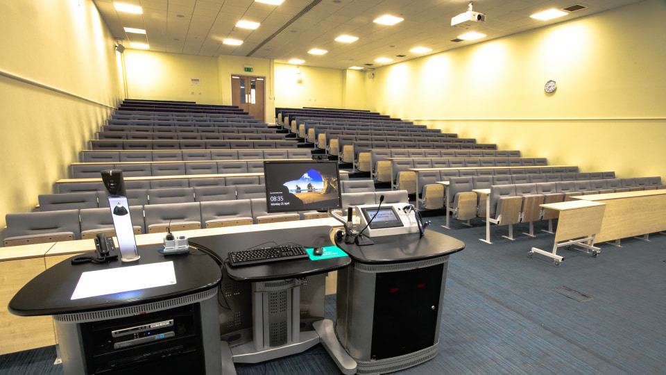 James France CC012 - Lecture hall with tiered seating, projector, and teaching podium equipped with computer and microphones.