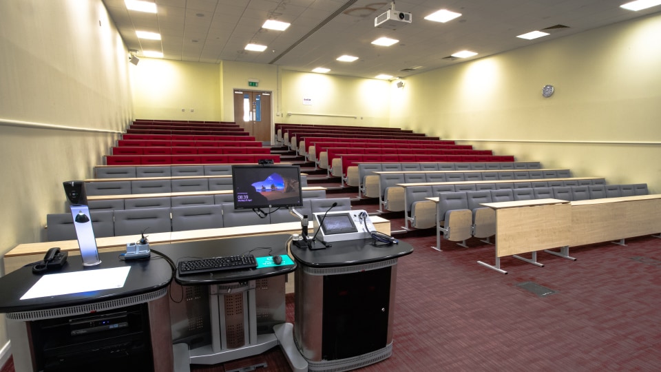 James France CC013 - Lecture hall with tiered red and gray seating, projector, and lectern equipped with computer and microphones.