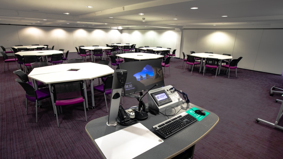 James France CC029a - Modern teaching room with hexagonal tables, purple chairs, and a presentation podium equipped with a computer and microphones.