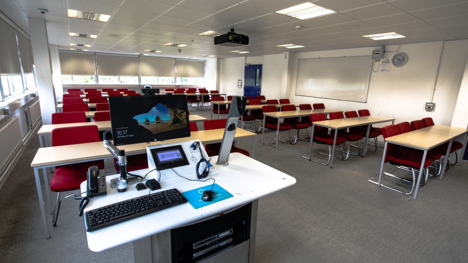 James France D109 - Teaching room with red chairs, wooden desks, projector, and instructor's podium with computer and phones.