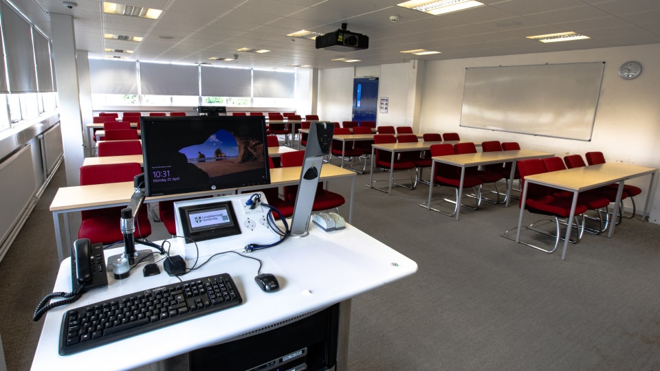 James France D201 - Modern teaching room with red chairs, tables, and a lectern equipped with computer and microphones. Large windows and whiteboard.