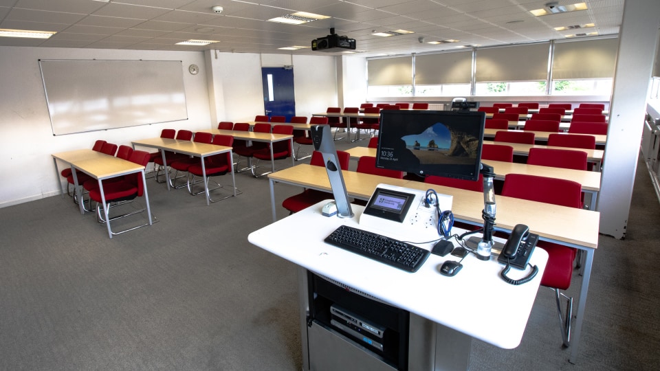 James France D202 - Teaching room with rows of red chairs, wooden desks, and a lectern featuring a computer and microphones.