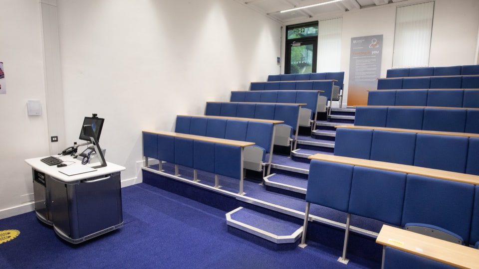 David Davies DAV030 - Lecture theatre with tiered blue seating, a lectern with computer and microphones, and a poster near the exit door.