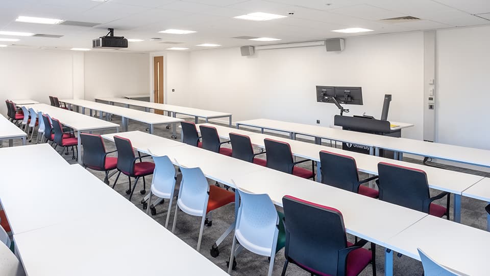 David Davies Dav1064 - Modern teaching room with long white tables, colorful chairs, dual monitors on a podium, and ceiling projector.