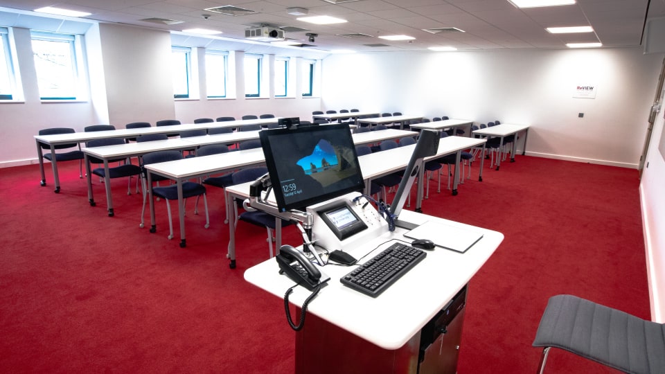 David Davies DAV1100 - Teaching room with red carpet, rows of tables and chairs, and a lectern with computer and phones. Bright windows.