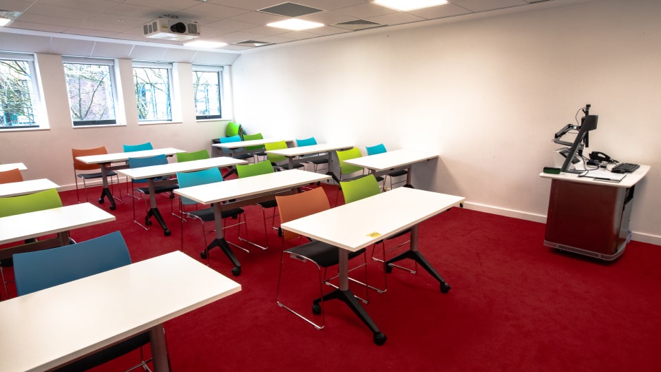 David Davies DAV1106 - Classroom with red carpet, colorful chairs, white desks, projector, and instructor's podium with computer near windows.