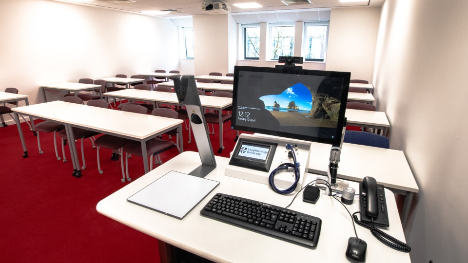 David Davies DAV1107 - Teaching room with red carpet, rows of desks and chairs. Front desk has computer, keyboard, phones, and cables.