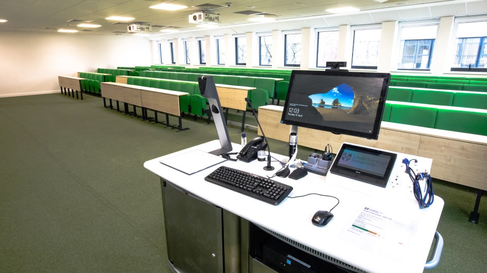 David Davies DAV1108 - Lecture theatre with green tiered seating, large windows, and a teaching podium equipped with computer, microphones, and control panel.