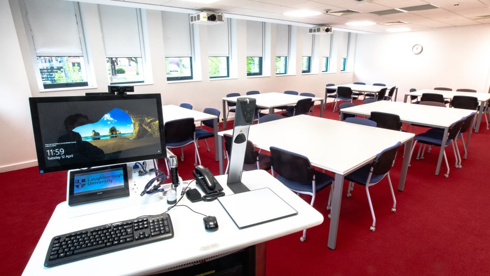 David Davies DAV1109 - Teaching room with red carpet, white tables, blue chairs, and a lectern with computer, keyboard, and phones. Windows line the wall.