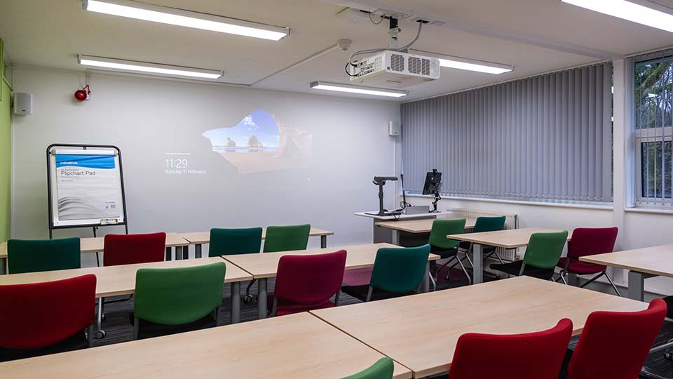 Teaching Room E007 - Classroom with colorful chairs, desks, projector, flipchart pad, and blinds. Projected screen displays time and date.