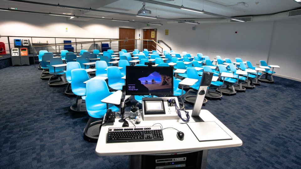 John Beckwith JB021 - Flat space with blue swivel chairs, desks, and a central teaching podium equipped with computer, microphones, and control panel.