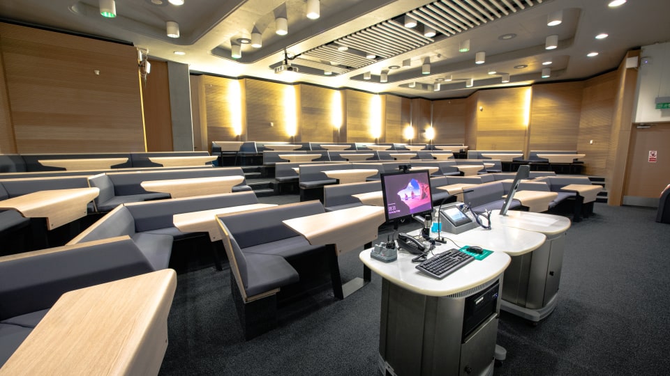 Design School LDS017 - Modern lecture theatre with tiered seating, wooden desks, and a central podium equipped with a computer and microphones.