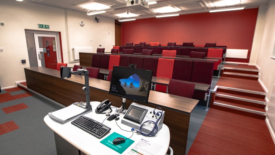 Frank Gibb RT040 - Lecture theatre with tiered seating, red and maroon chairs, a lecturer's desk with computer, keyboard, phones, and projector controls.