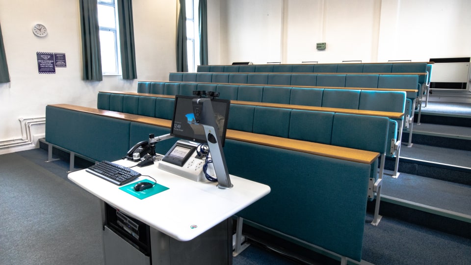 Schofield SCH105 - Lecture theatre with tiered seating, teal upholstery, and a lecturer's podium equipped with a computer, keyboard, and microphones.