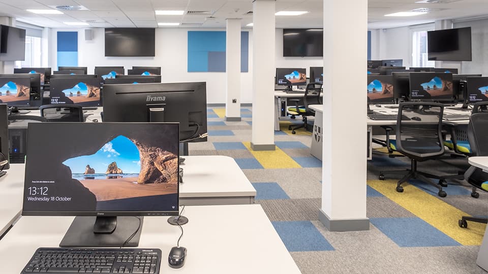 Teaching Room WAV001 - Spacious computer lab with rows of desks, monitors displaying a beach scene, ergonomic chairs, and colorful carpet tiles.