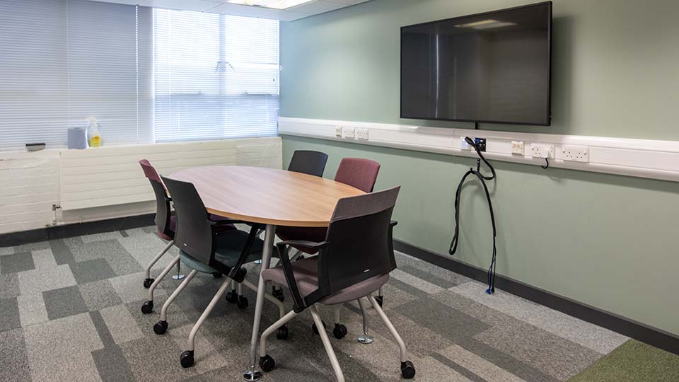 Teaching Room WAV008 - Teaching room with oval table, five chairs, wall-mounted TV, power outlets, and window blinds. Neutral colors, carpeted floor.