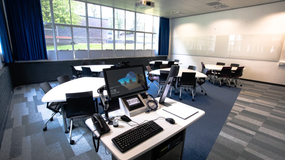 Wavy Top WAV027 - Modern teaching room with hexagonal tables, chairs, large windows, blue curtains, and a presentation desk equipped with computer and phones.