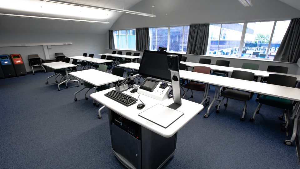 Wavy Top WAV046 - Teaching room with a podium, computer, and microphones. Rows of tables and chairs face large windows with curtains.