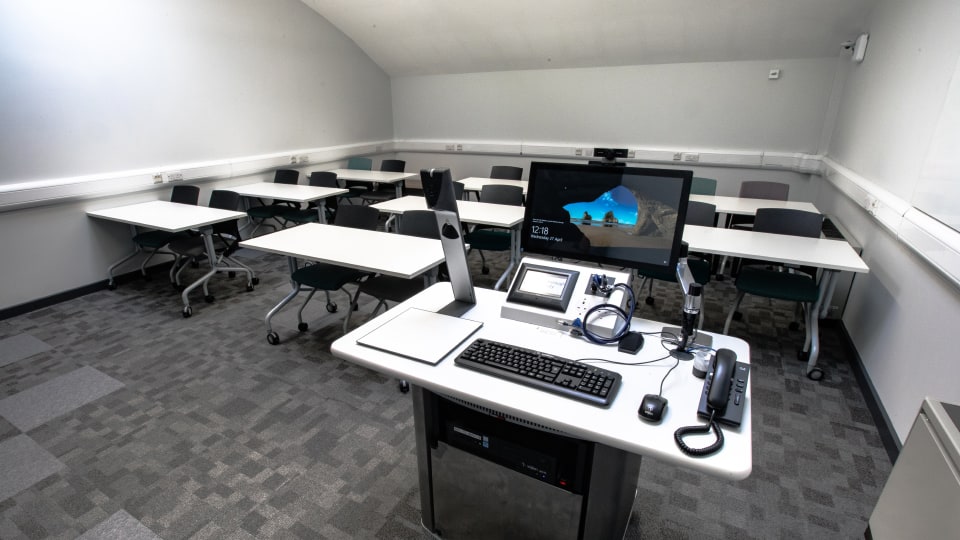 Wavy Top WAV116 - Teaching room with a central lectern featuring a computer, keyboard, microphones, and phones. Rows of tables and chairs fill the space.