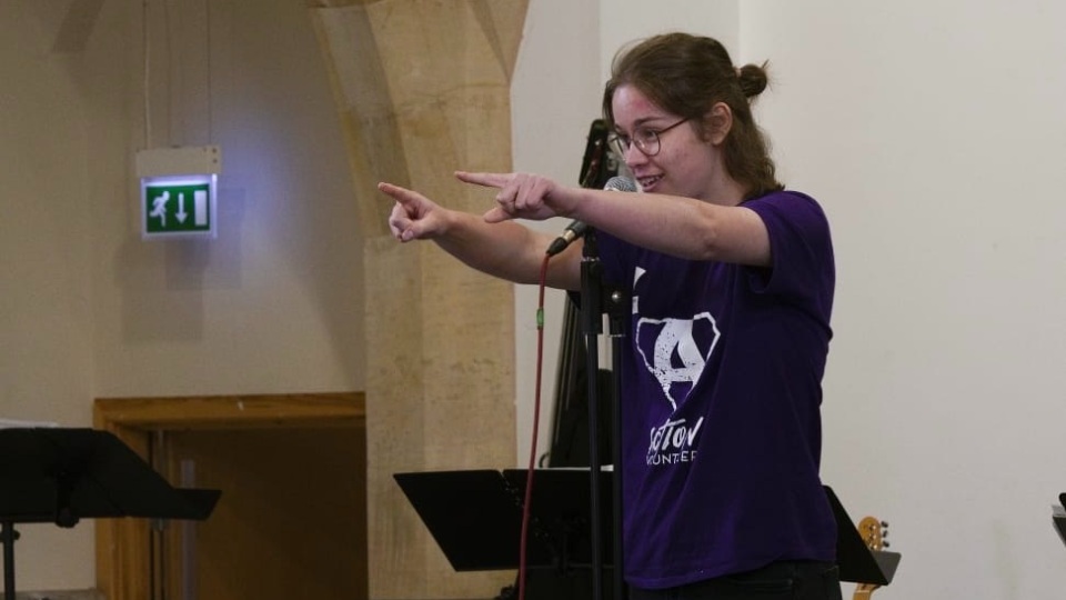 Daisy Newton wearing an Action t-shirt and pointing forwards whilst speaking into a microphones.